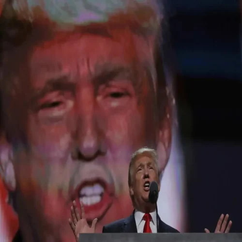 Donald Trump at a podium with a huge screen of his face behind him.
